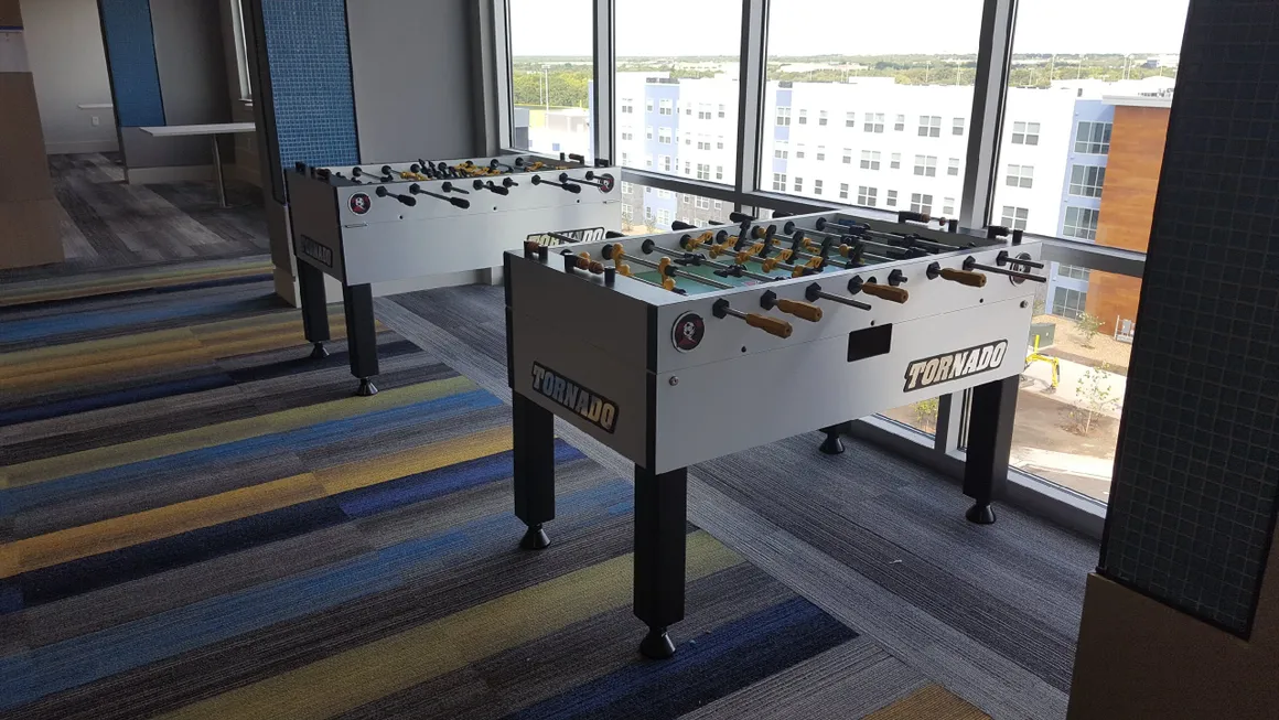 Two Item # MO17001-FB foosball tables in a room with windows.