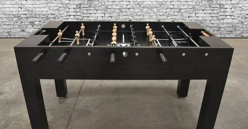 A black foosball table (Item # MO17001-FB) with wooden sticks and a brick wall.