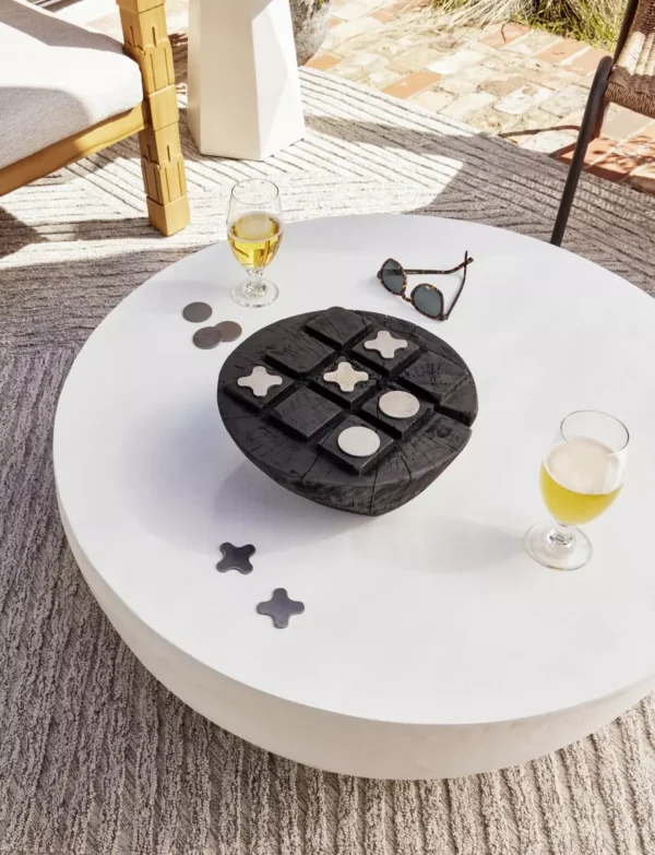 A round white FH25031-AC table with a black tic-tac-toe game, two glasses of white wine, sunglasses, and pebbles, in a sunny patio setting.