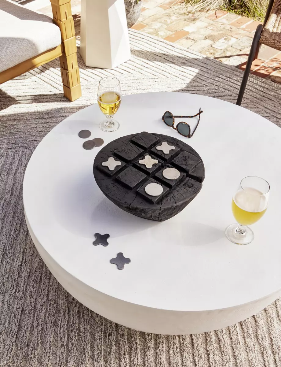 A round white FH25031-AC table with a black tic-tac-toe game, two glasses of white wine, sunglasses, and pebbles, in a sunny patio setting.