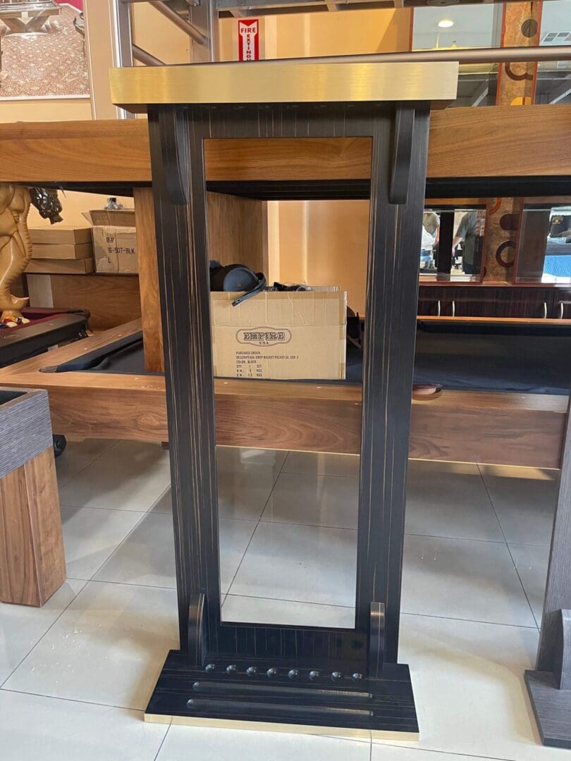 A tall, black wooden pedestal with a metal top (Item # PH22030-CRW), displayed in a shop with cluttered shelves and boxes in the background.